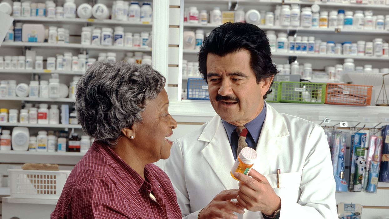 A woman in red speaks to a man in a white coat who holds a yellow pill bottle.