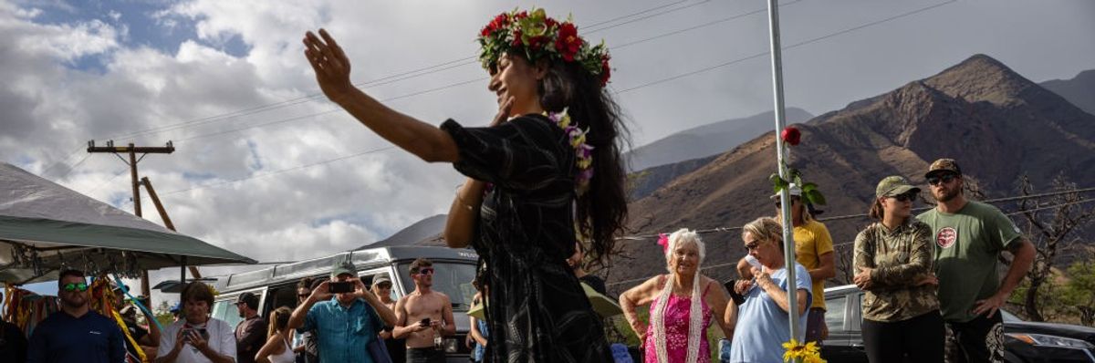 A woman perfomrs a hula dance in front of a crowd of mourners. 