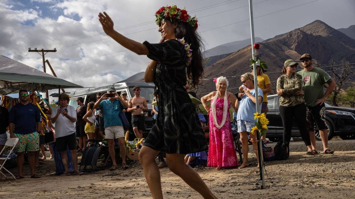 A woman perfomrs a hula dance in front of a crowd of mourners.