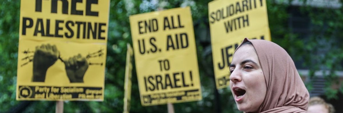 A woman shouts and shakes a tambourine in front of Palestine solidarity flyers in yellow and black.