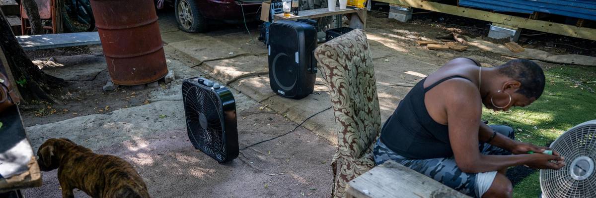 A woman sits next to a fan outside her home.