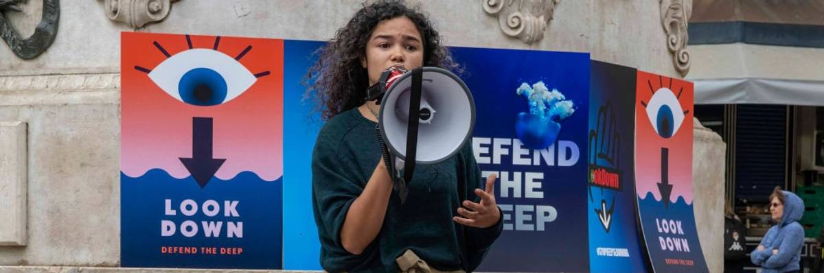 A woman stands with a megaphone in front of posters saying, "Look Down," and "Defend the Deep."
