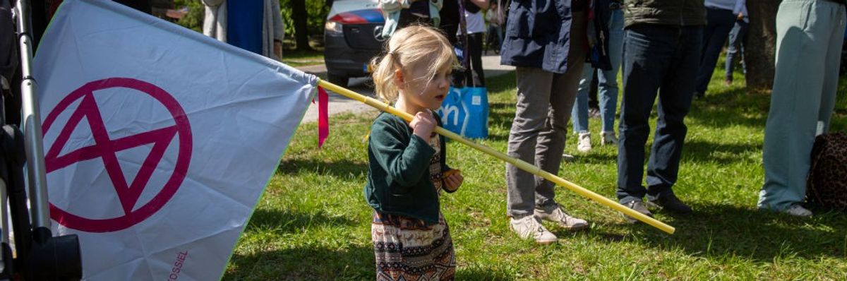 A young child standing on grass holds a red and white Extinction Rebellion flag.