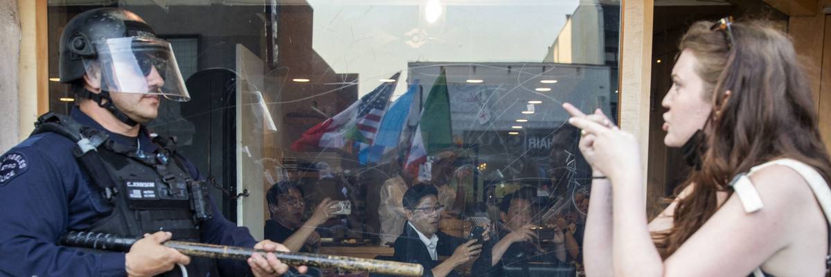 A young female-appearing protester and police officer in riot gear face each other.