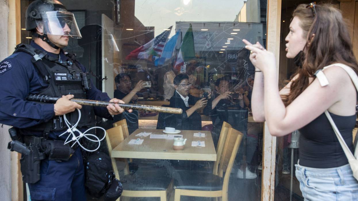 A young female-appearing protester and police officer in riot gear face each other.
