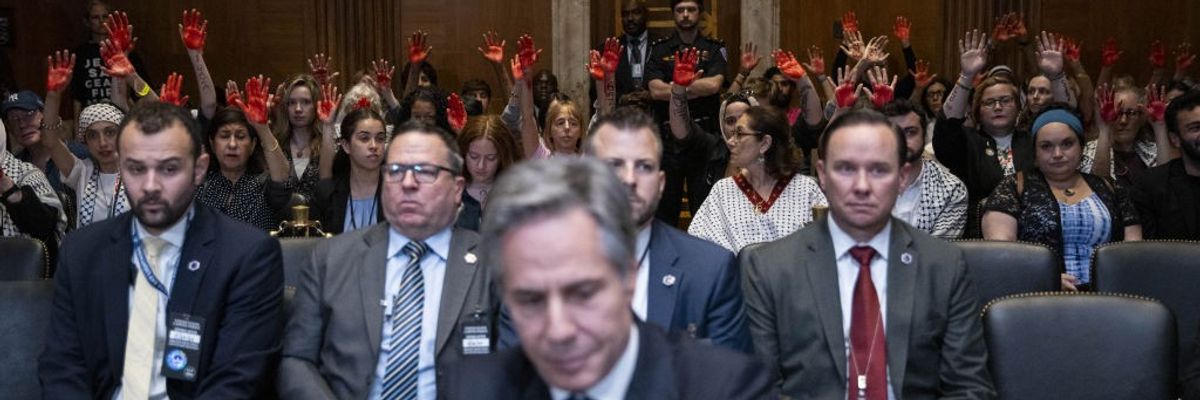 Activists raise red hands behind Antony Blinken.