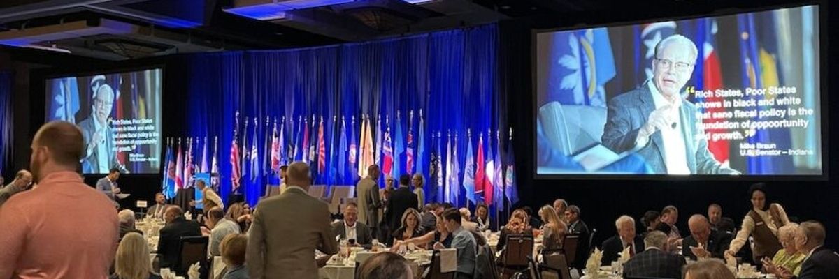 ALEC members sit at tables watching a video on a screen.