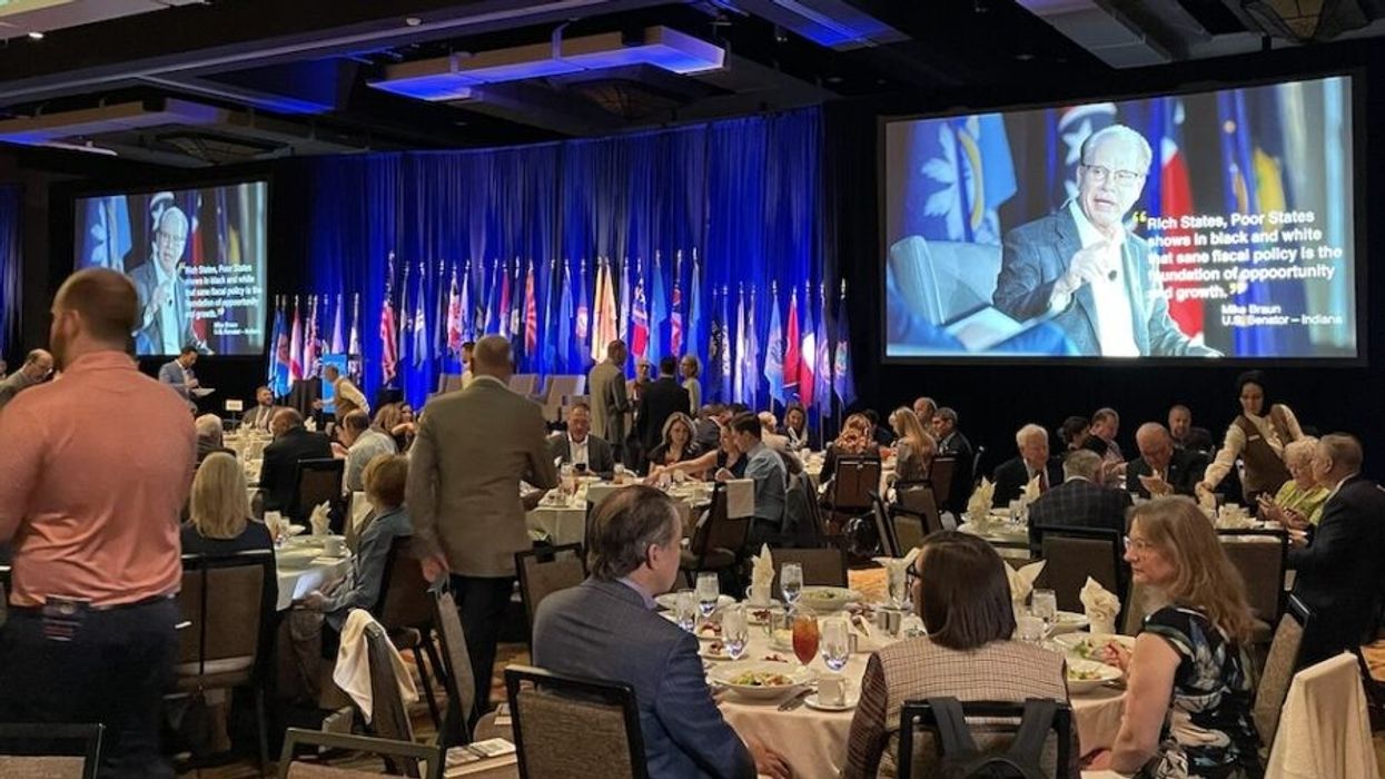 ALEC members sit at tables watching a video on a screen.