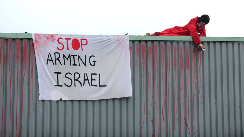 An activist in red lies on a roof next to a white banner reading, "Stop arming Israel."