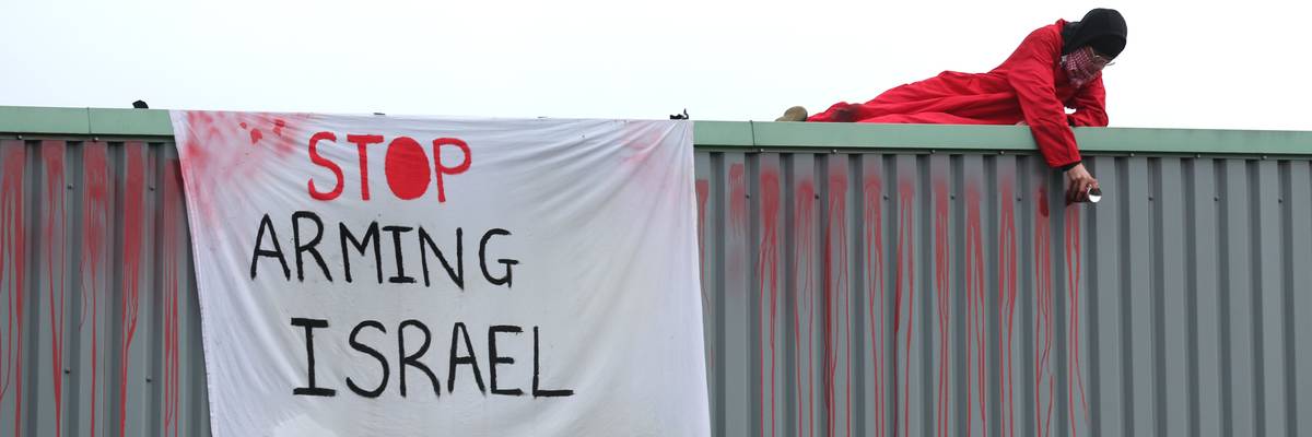 An activist in red lies on a roof next to a white banner reading, "Stop arming Israel."