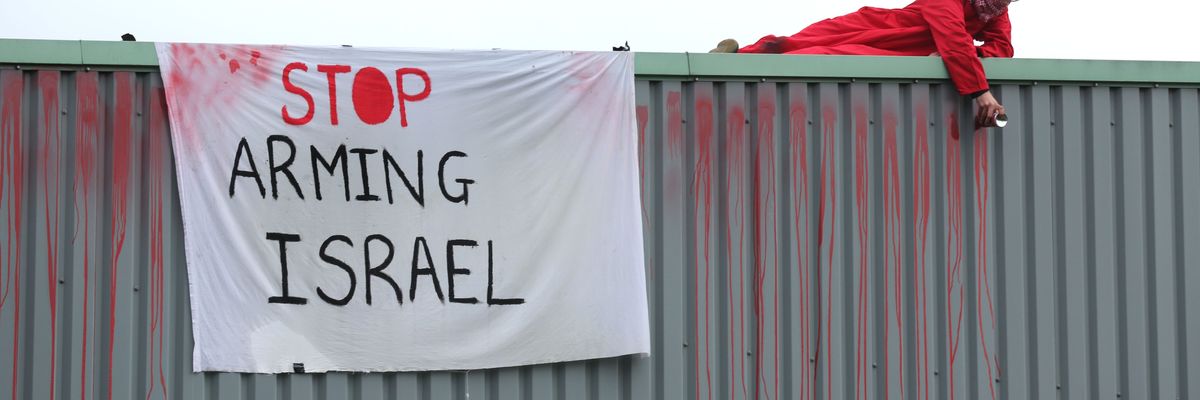 An activist in red lies on a roof next to a white banner reading, "Stop arming Israel."