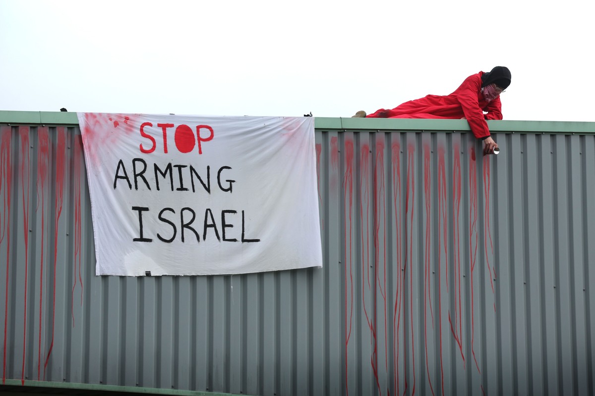 An activist in red lies on a roof next to a white banner reading, "Stop arming Israel."