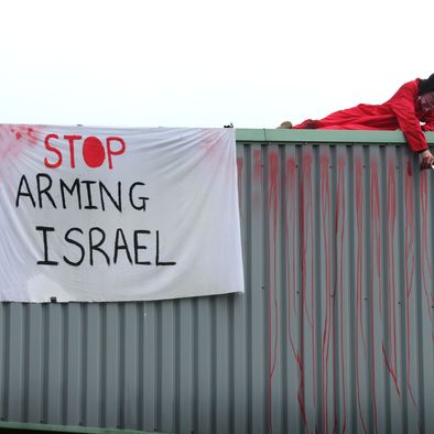 An activist in red lies on a roof next to a white banner reading, "Stop arming Israel."