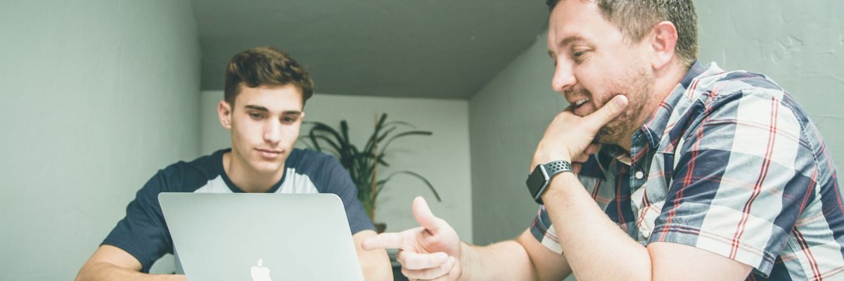 An older man points at a laptop while a younger man looks on.