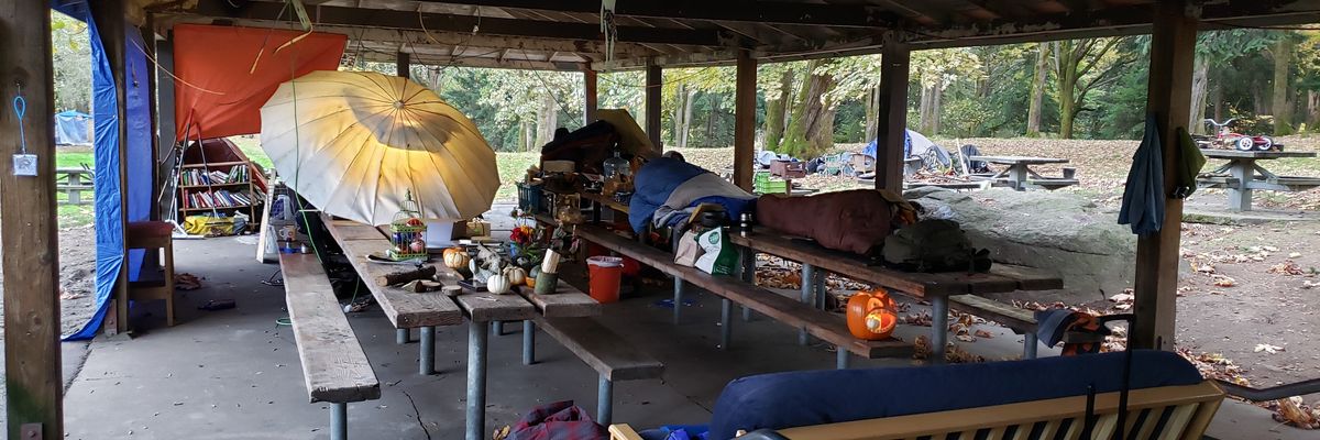 An unhoused person sleeps on a picnic table in a park shelter.