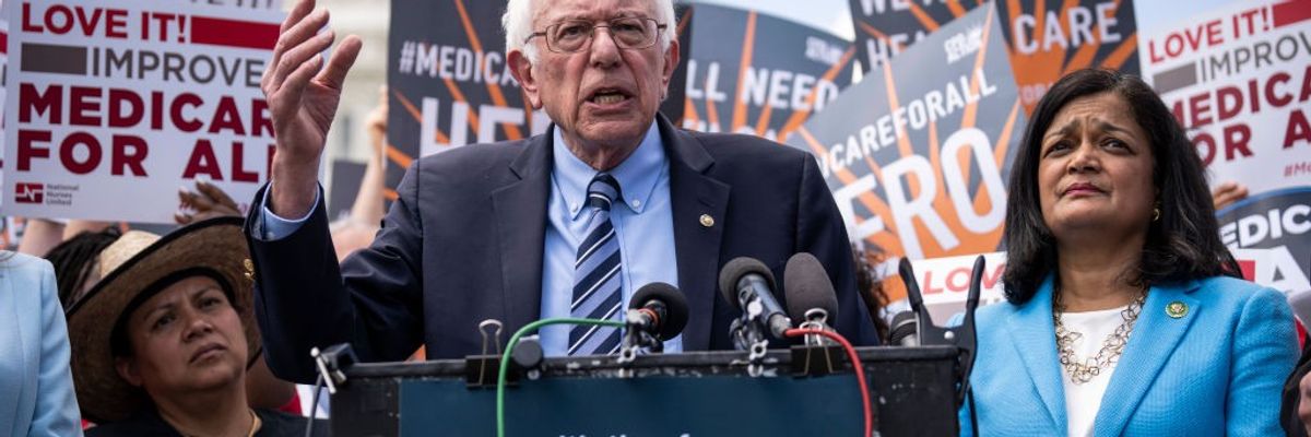 Bernie Sanders and Pramila Jayapal stand at a podium in front of people supporting Medicare for All.