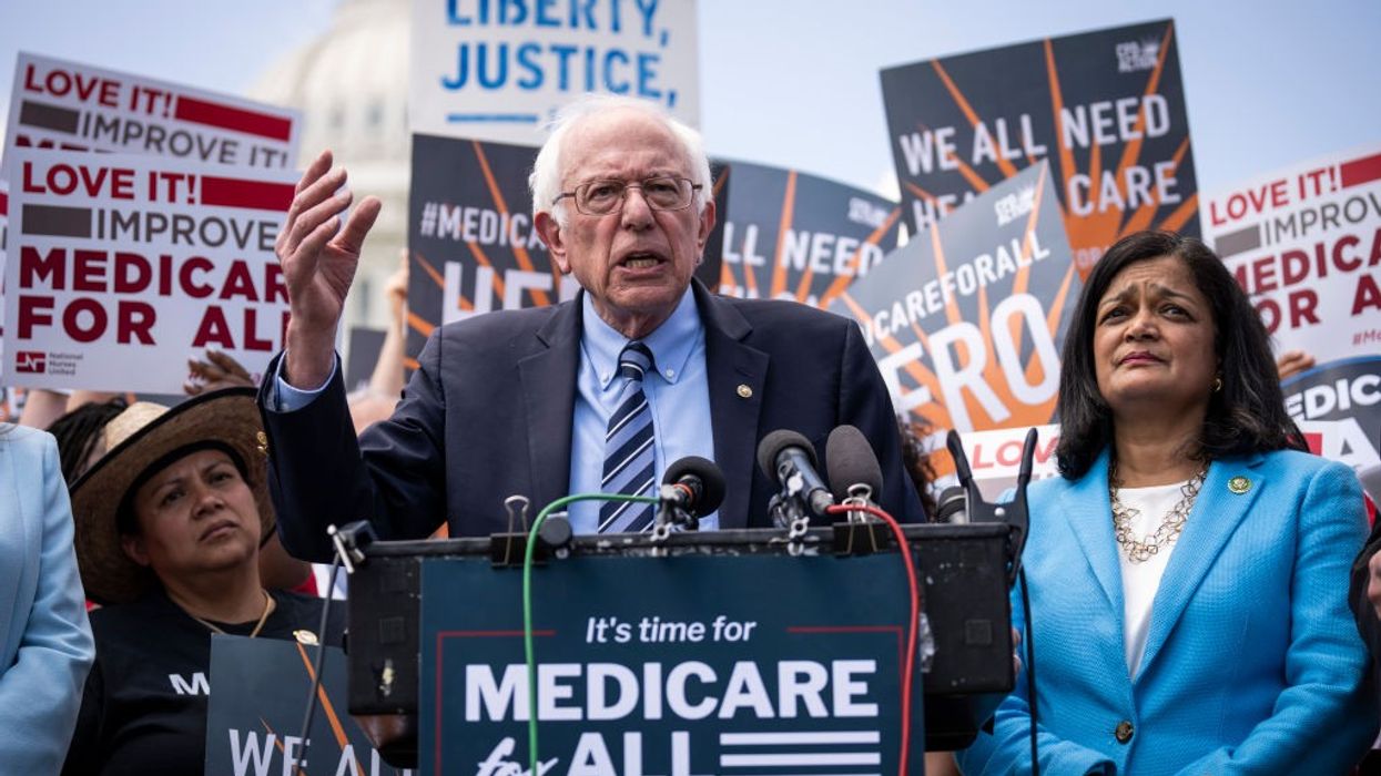 Bernie Sanders and Pramila Jayapal stand at a podium in front of people supporting Medicare for All.