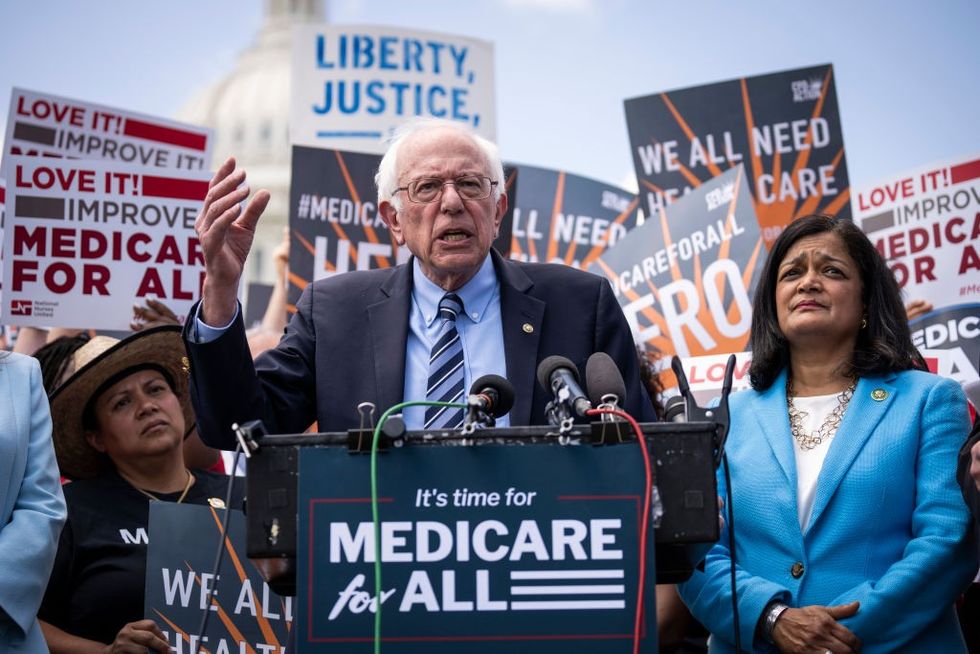 Bernie Sanders and Pramila Jayapal stand at a podium in front of people supporting Medicare for All.