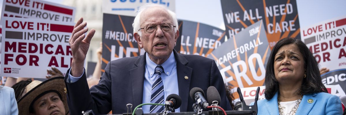 Bernie Sanders and Pramila Jayapal stand at a podium in front of signs.