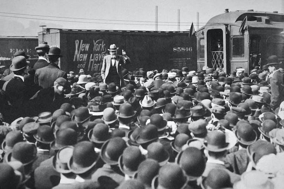 Black-and-white image of a man speaking from a train to a crowd of men in top hats