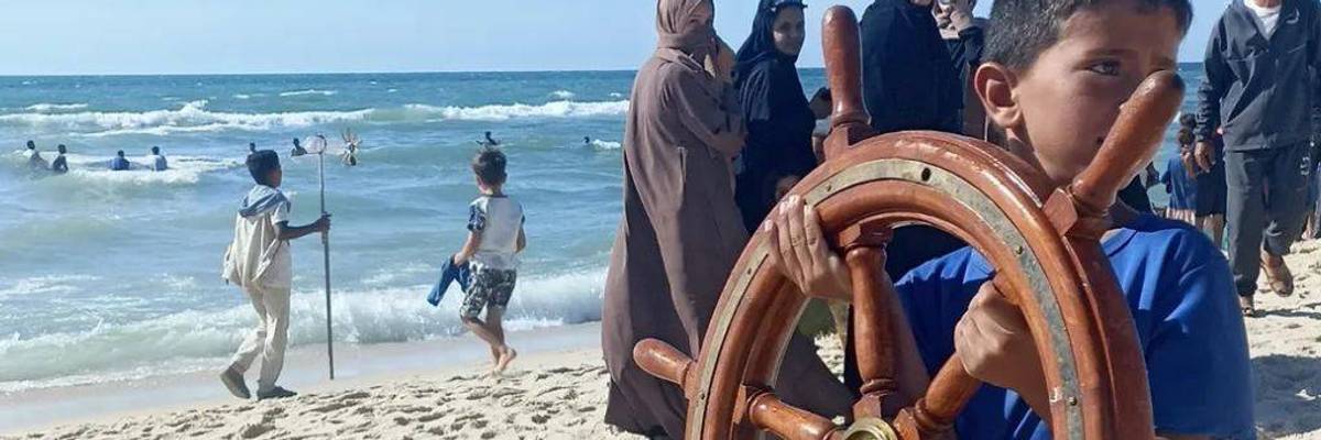 Child in Gaza carries sailing ship steering wheel onto beach.