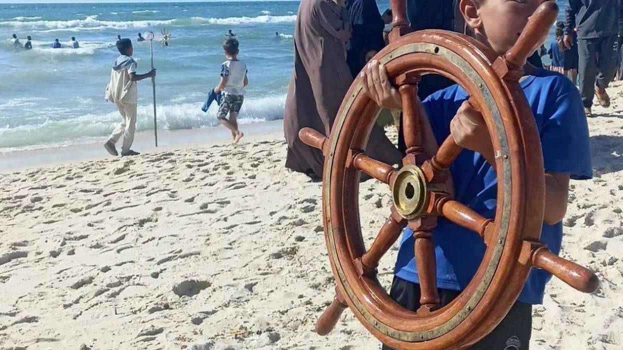 Child in Gaza carries sailing ship steering wheel onto beach.