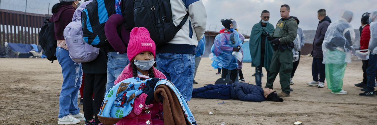 child waits with other migrants for transport as an ill man is tended to
