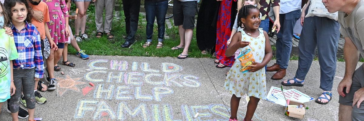 Children and parents stand next to a message in sidewalk chalk reading, "Child checks help families thrive."
