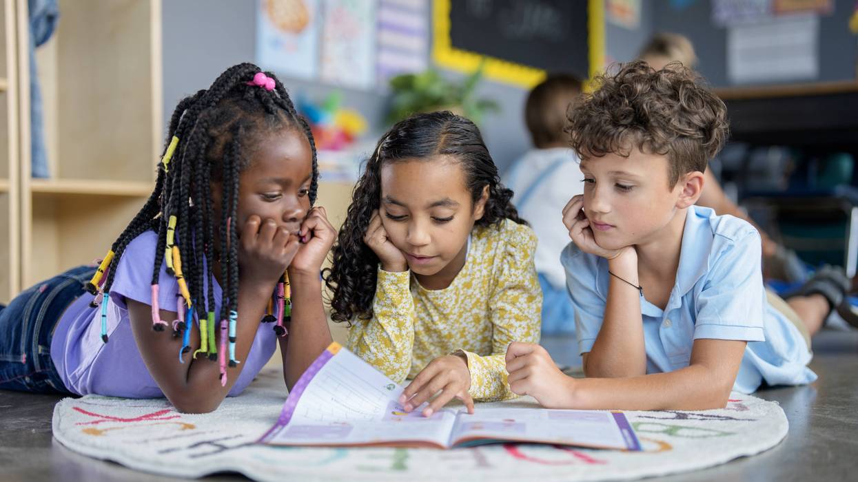 Children lie on a rug in a classroom, reading a book.