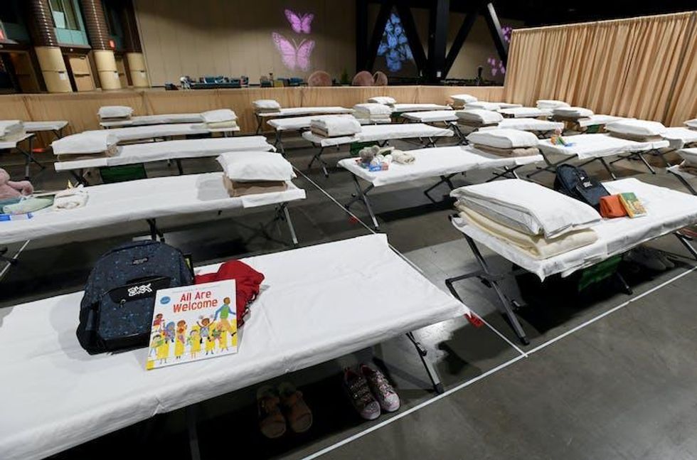 Cots lined up in a large space, some with backpacks and children's books on them