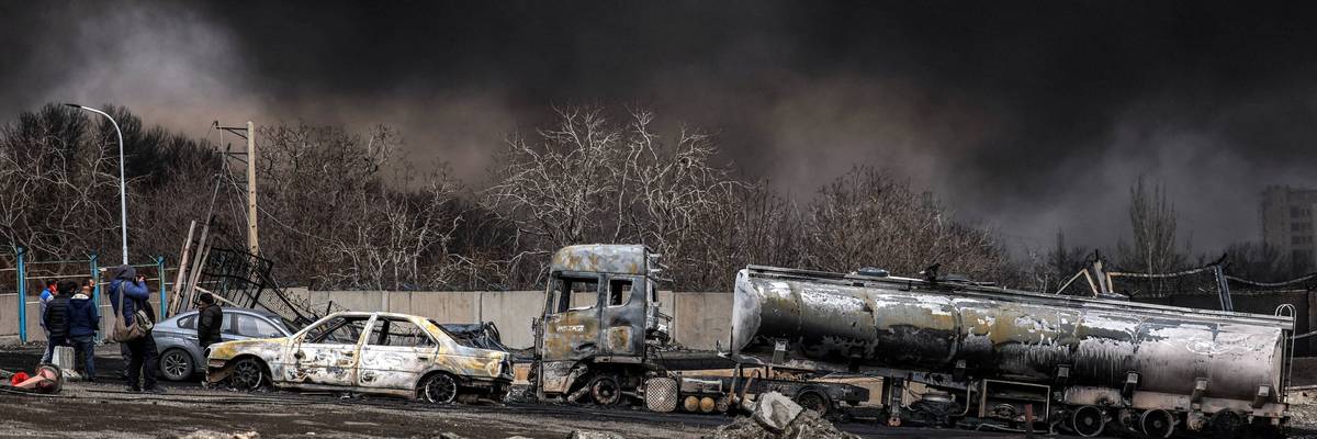 Destroyed vehicles in front of a dark cloud from US strikes on Iran.