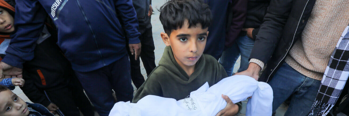 During prayers for the dead outside a Gaza hospital, a young boy holds the shrouded body of his infant sibling killed in an Israeli air strike