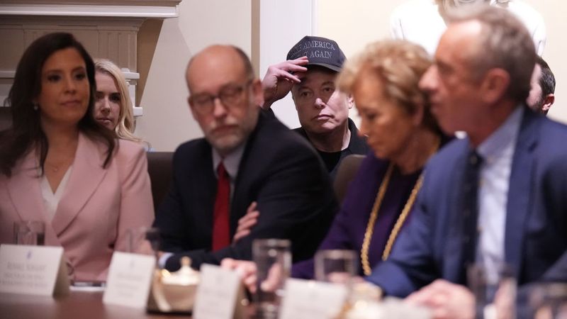 Elon Musk, framed by Russell Vought and Linda McMahon