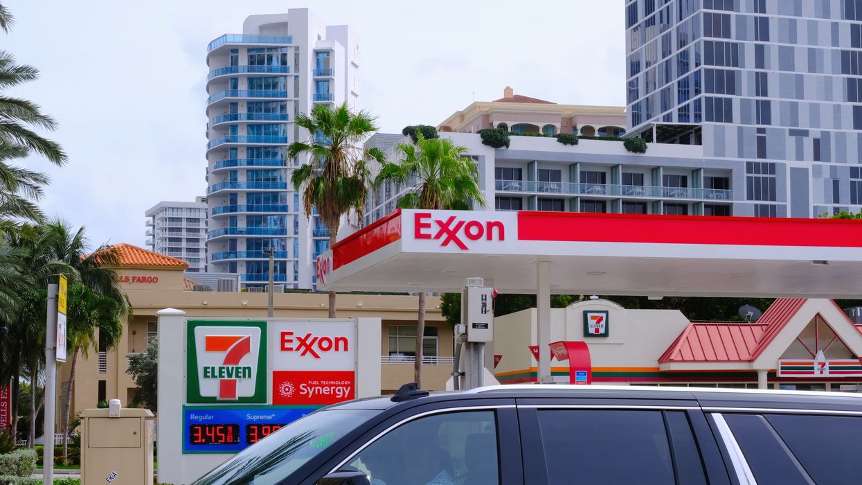 Exxon gas station with 7-Eleven branding is seen against modern high-rise buildings, palm trees, and a passing black SUV.
