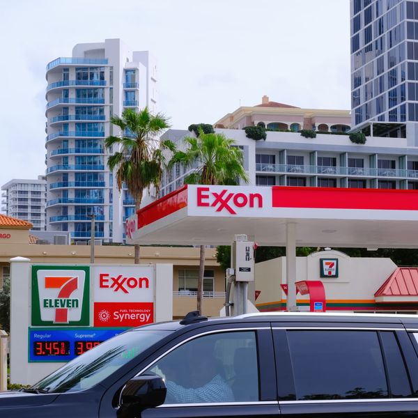 Exxon gas station with 7-Eleven branding is seen against modern high-rise buildings, palm trees, and a passing black SUV.