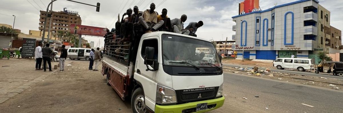 Figures ride on top of a van down a litter-strewn street.