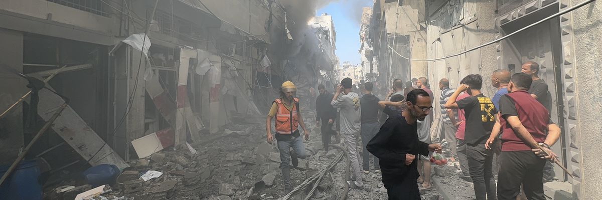 firefighter and a small crowd of people stand in a destroyed street and look at building that has smoke coming out of it.