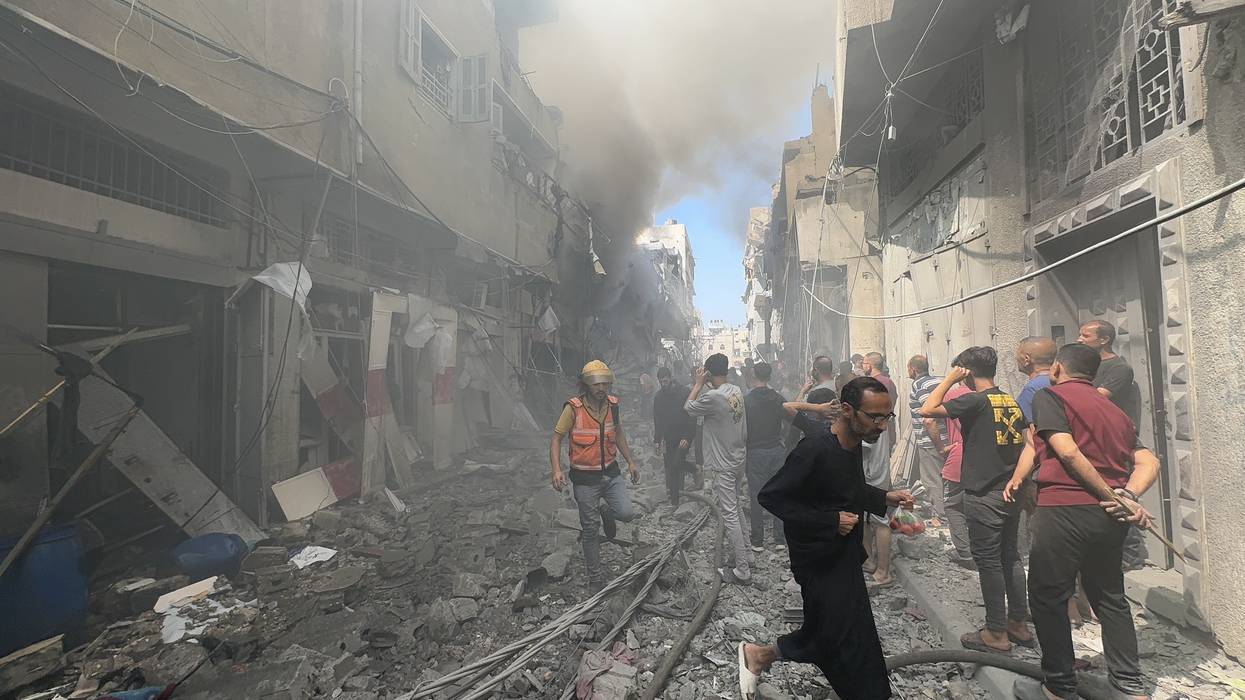 firefighter and a small crowd of people stand in a destroyed street and look at building that has smoke coming out of it.