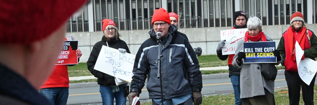 ICU nurse speaks into a microphone. protestors with signs stand behind him.