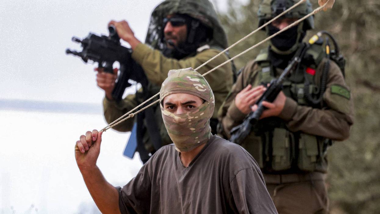 Israeli soldiers stand with rifles pointed as a masked settler swings a slingshot with rock loaded