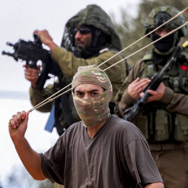 Israeli soldiers stand with rifles pointed as a masked settler swings a slingshot with rock loaded