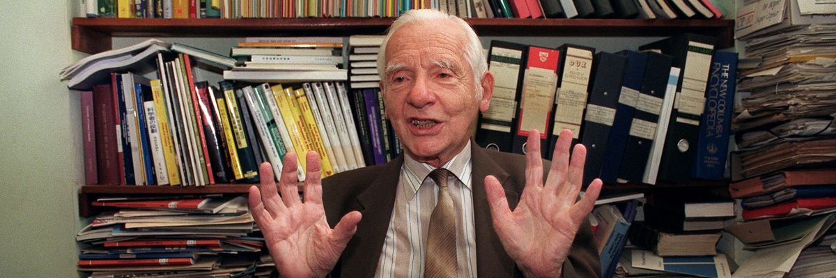Joseph Rotblat with his hands raised sitting in front of books.