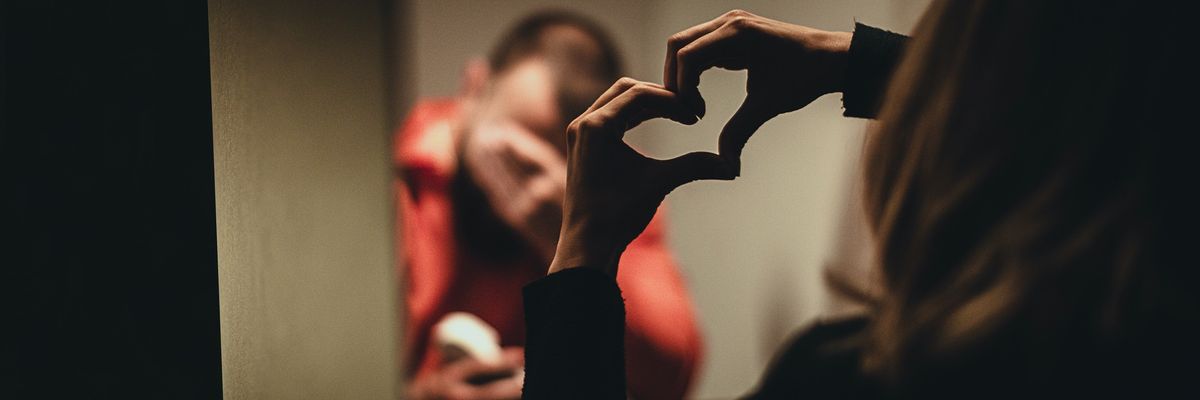 Man and woman visit in prison phone booth as she holds up "heart" sign