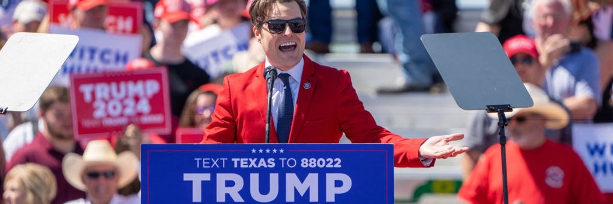 Matt Gaetz addresses a Trump rally wearing a red suit, white shirt, blue tie, and and sunglasses