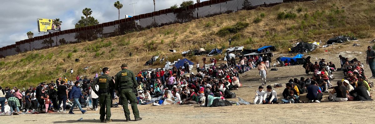 Migrants cluster in an open-air detention site while U.S. border officials look on