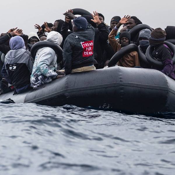 Migrants onboard a rubber boat wave and gesture as they wait to be rescued