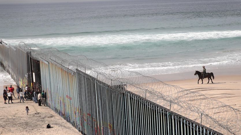 migrants stand on a beach by the border wall