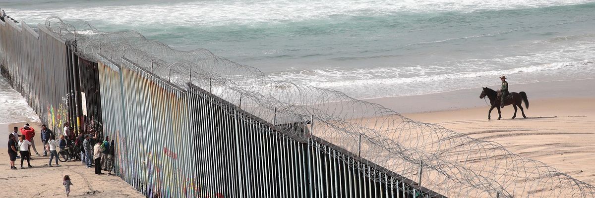 migrants stand on a beach by the border wall