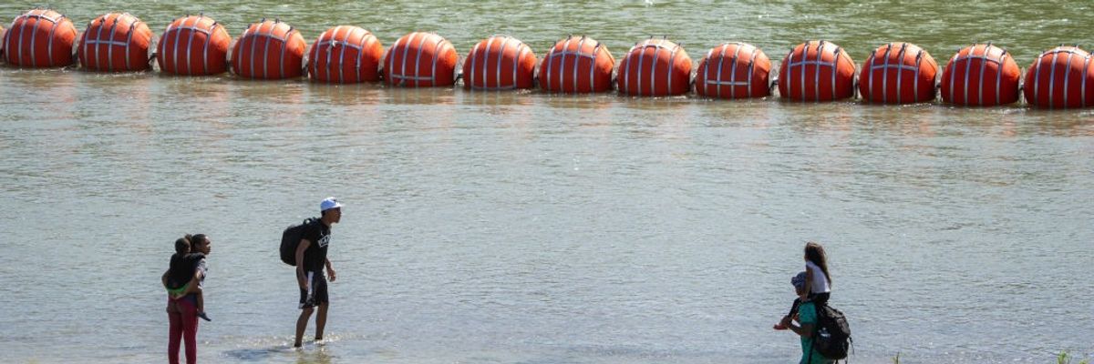 Migrants walk by a floating barrier along the Rio Grande