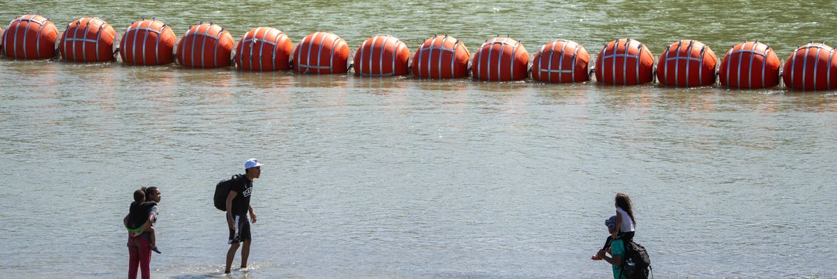Migrants walk by a floating barrier along the Rio Grande